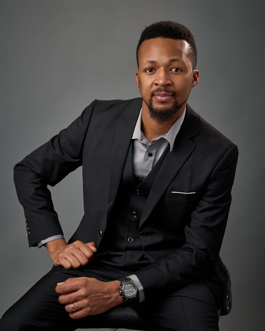 Portrait of Richard Abiona in a black three-piece suit, seated against a grey backdrop